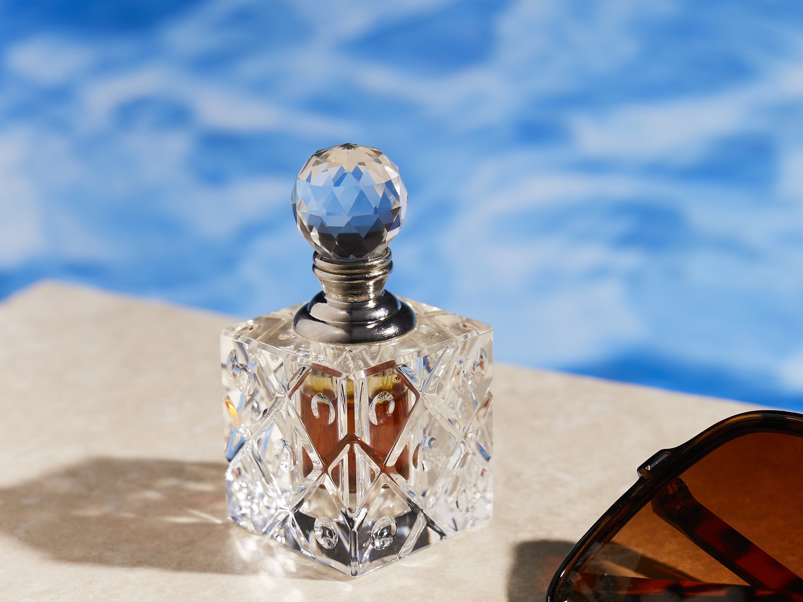 Crystal oud oil bottle poolside with sunglasses under a blue sky